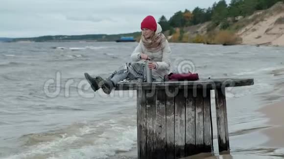 年轻女子穿着温暖的衣服坐在海边坐在木盘管上喝着热水瓶里的热茶寒冷的天气暴风雨视频的预览图