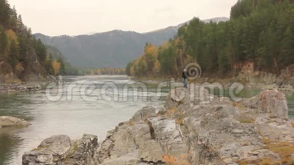 山河在岩石之间流淌着松树旅行者站在河边的一块大石头上视频的预览图