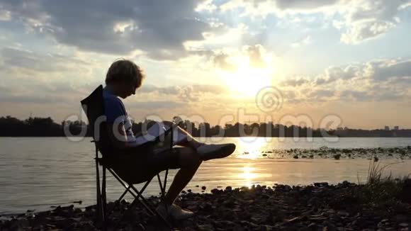金发碧眼的人在日落的河岸上阅读并写下他的笔记视频的预览图