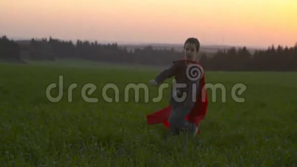 日落时穿着超级英雄斗篷的男孩在田野里奔跑视频的预览图