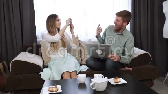 情侣坐在家里的沙发上做智能手机上的自拍照片看着笔记本电脑屏幕微笑视频的预览图