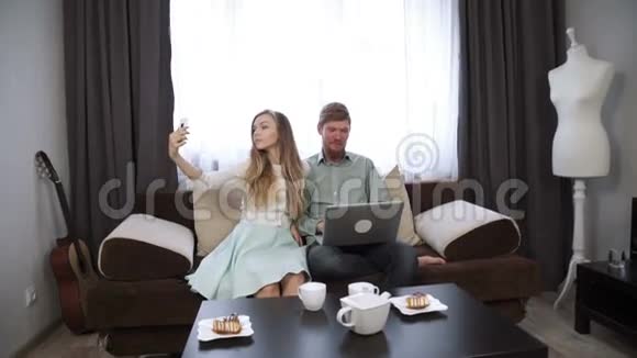 情侣坐在家里的沙发上在智能手机上自拍看着笔记本电脑屏幕微笑视频的预览图