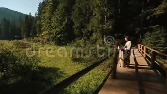 在喀尔巴阡山的木桥上穿着传统的乌克兰服装的幸福美丽的一对夫妇轻轻地接吻玩得开心视频的预览图