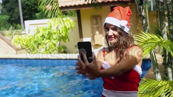在阳光明媚的日子里年轻的美女戴着圣诞老人的帽子在游泳池里用手机做自我画像视频的预览图