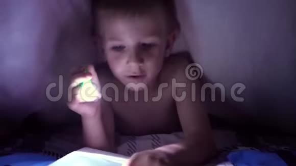 一个孩子晚上用手电筒在毯子下面读一本书有着淡发和蓝眼睛的男孩视频的预览图