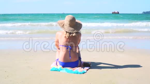 年轻的金发女人带着蓝色比基尼和草帽坐在海滩上看着大海视频的预览图