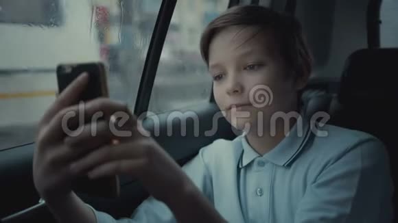 不幸的是不幸的小男孩在雨天开车穿过城市在智能手机上使用社交网络视频的预览图