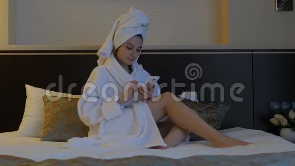 一个穿着白色外套的年轻漂亮的女人坐在酒店卧室的床上在电话里写短信视频的预览图