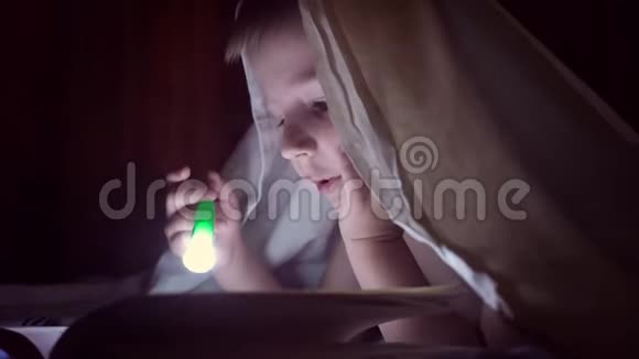 一个孩子晚上用手电筒在毯子下面读一本书有着淡发和蓝眼睛的男孩视频的预览图