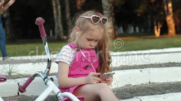 一个穿着粉红色裙子的小女孩坐在台阶上的公园里戴着耳机享受着智能手机视频的预览图