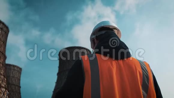专业工程师戴着白色头盔和夹克看着工厂的管道冷却塔上有白色的视频的预览图