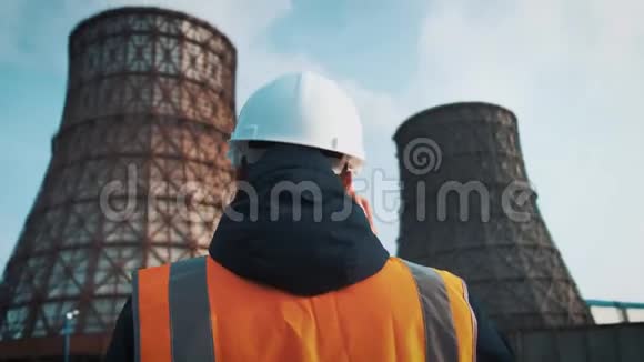 专业工程师戴着白色头盔和夹克看着工厂的管道冷却塔上有白色的视频的预览图