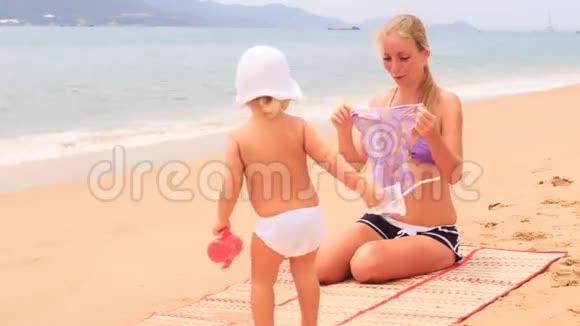 妈妈给坐在沙滩上的女儿看泳衣视频的预览图