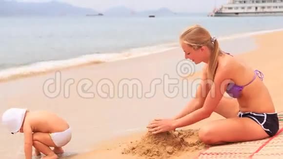 年轻的母亲坐在垫子上靠船建造沙堡视频的预览图