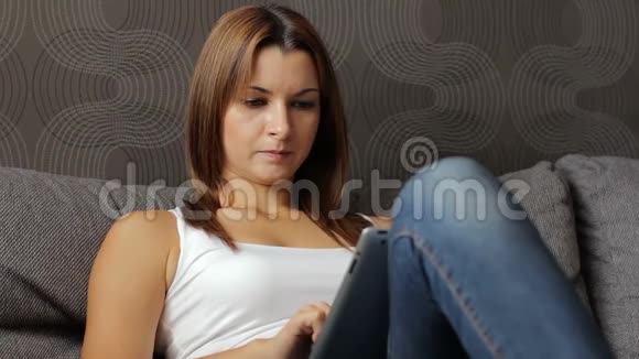 年轻女性看数字平板电脑的肖像有平板电脑的女人坐在沙发上使用平板电脑的女人视频的预览图
