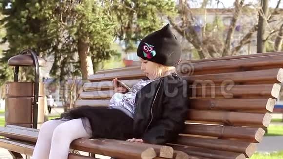 一个女孩在晴天坐在长凳上用手机在公园里的肖像视频的预览图