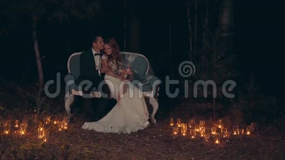 新婚夫妇在夜晚拥抱和亲吻坐在黑暗的森林里的沙发上视频的预览图
