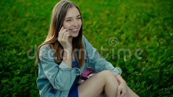 年轻漂亮的女人坐在夏季城市公园的草地上用手机聊天视频的预览图