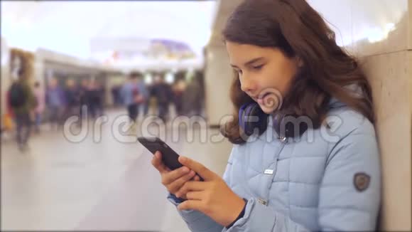 十几岁的女孩带着智能手机和耳机等待地铁很多人挤在地下交叉路口地铁地下视频的预览图