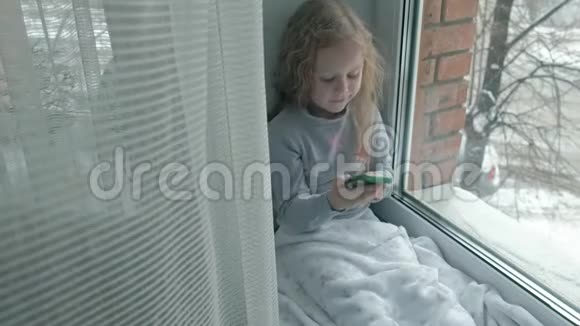 幸福的小女孩留着红色波浪发坐在窗台上盖着毯子用电话聊天视频视频的预览图