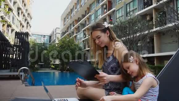 年轻的母亲在游泳池附近用笔记本电脑度假小女儿在慢动作视频的预览图