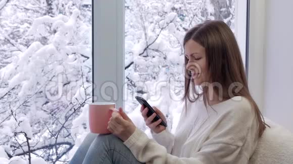 漂亮的女孩坐在窗台上喝茶用智能手机冬天在外面视频的预览图