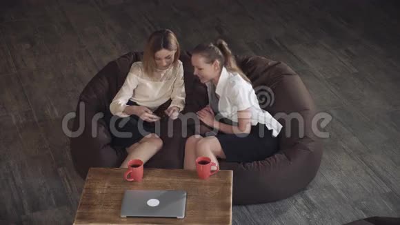 两位迷人的商务女性在沙发上放松在电话里看照片视频的预览图