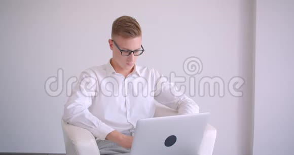 年轻英俊的白种人商人戴着眼镜手持笔记本电脑坐在室内的扶手椅上视频的预览图