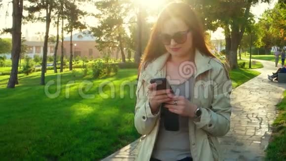 美丽的年轻红发女子的春日画像戴着滑稽的太阳镜站在公园的背光下发短信视频的预览图