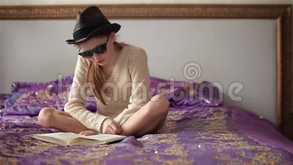 戴太阳镜戴帽子的小女孩坐在床上看书视频的预览图