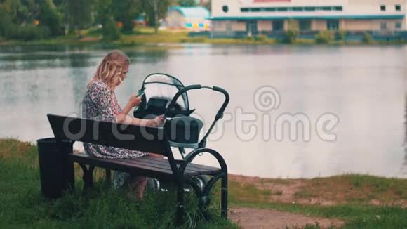母亲摇摆婴儿车和使用智能手机在夏季公园湖视频的预览图