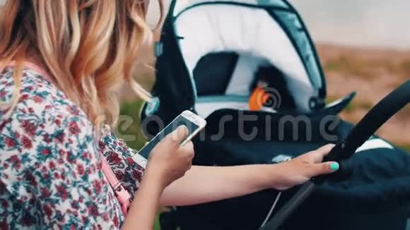 母亲摇摆婴儿车和使用智能手机在公园夏日阳光明媚视频的预览图