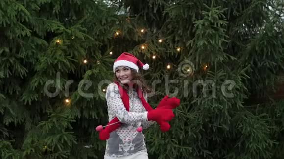 戴着红领巾和手套的圣诞老人帽子的快乐的女人在圣诞树的背景下跳舞视频的预览图