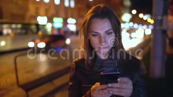 迷人的女人在夜城的街道上行走时使用智能手机视频的预览图