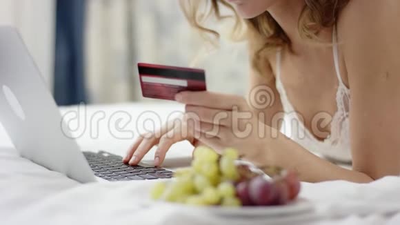 穿着睡衣的漂亮女人躺在床上使用她的笔记本电脑和信用卡视频的预览图