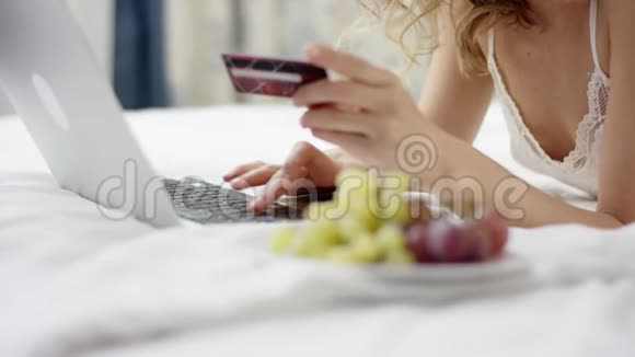 穿着睡衣的女人躺在床上用她的笔记本电脑和信用卡进行网上银行业务视频的预览图