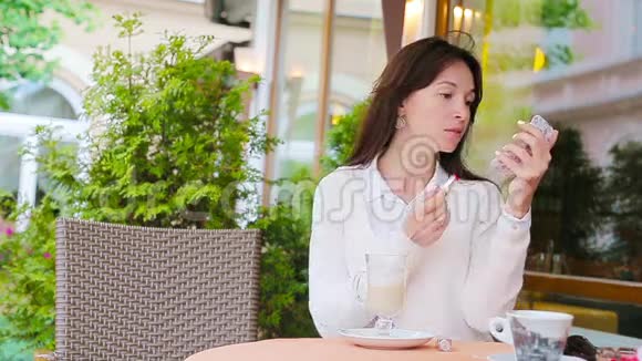 漂亮的女人坐在户外咖啡馆里用红色的口红画她的嘴唇年轻女孩带着美味的卡布奇诺视频的预览图