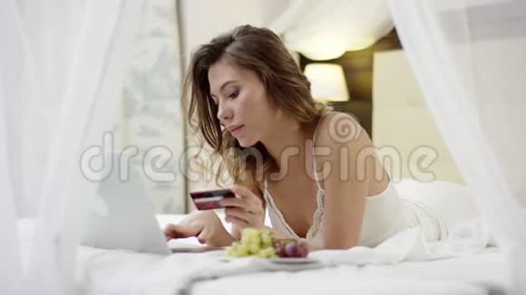 穿着睡衣的漂亮女人躺在床上用她的笔记本电脑和信用卡视频的预览图