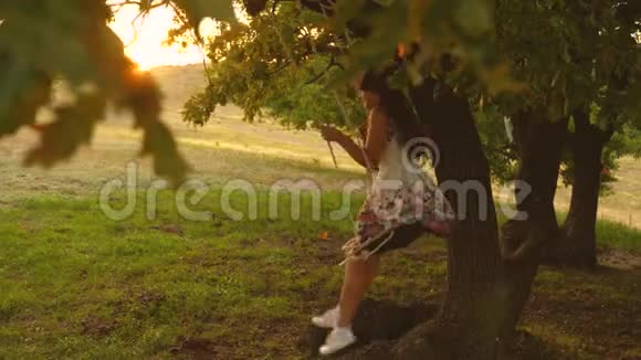 美丽的女孩在橡木树枝上的绳索秋千上摆动公园里穿着白色连衣裙的年轻女孩少女喜欢视频的预览图
