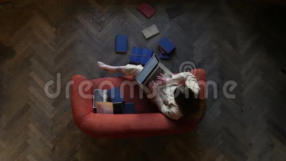 情绪化的年轻女人在沙发上用现代笔记本电脑发短信开心地笑着被地板上的许多书包围着视频的预览图