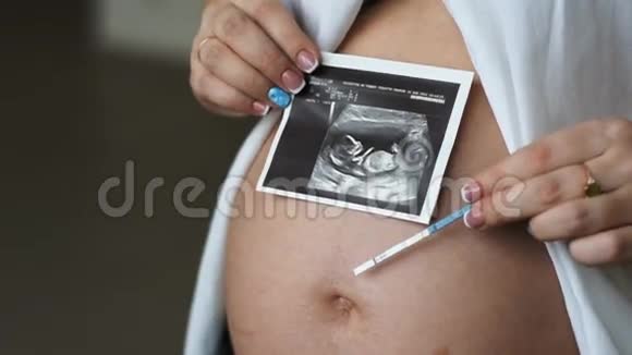 一位怀孕的年轻妇女在腹部拿着一张照片和一个怀孕测试视频的预览图