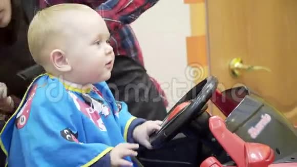 小男孩剪理发师他坐在一张看起来像汽车的椅子上妈妈它分散注意力和他玩视频的预览图