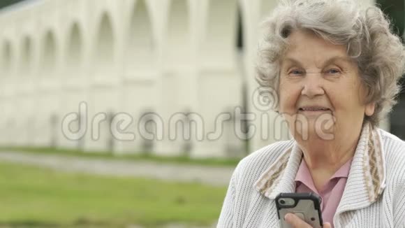 微笑的老妇人在户外手持银色智能手机视频的预览图
