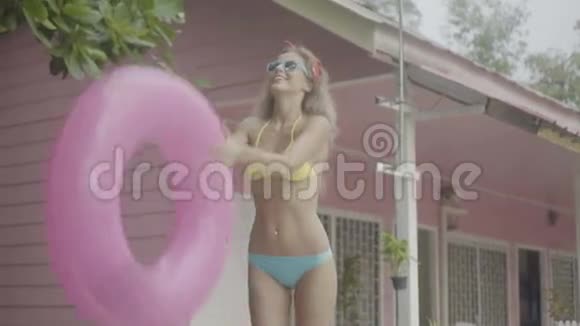 穿着比基尼戴着墨镜戴着粉色充气戒指的漂亮年轻幸福的女人在粉色房子的背景下玩视频的预览图