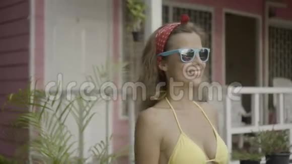 穿着比基尼和太阳眼镜的漂亮年轻快乐的女人在粉色房子的背景下孤独地行走视频的预览图