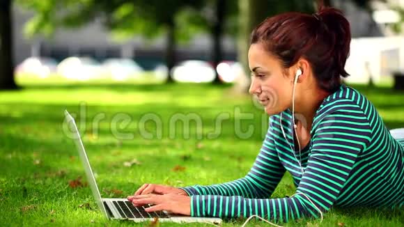 漂亮的学生躺在草地上用手提电脑听音乐视频的预览图