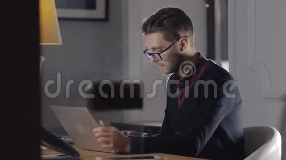 多任务处理英俊的年轻人戴着眼镜坐在现代的办公室里和Laptop一起工作自由职业者视频的预览图