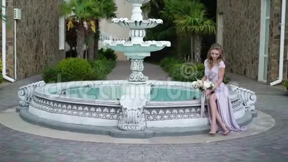 年轻的金发女郎穿着内衣在喷泉附近的花园里摆着新娘花束视频的预览图
