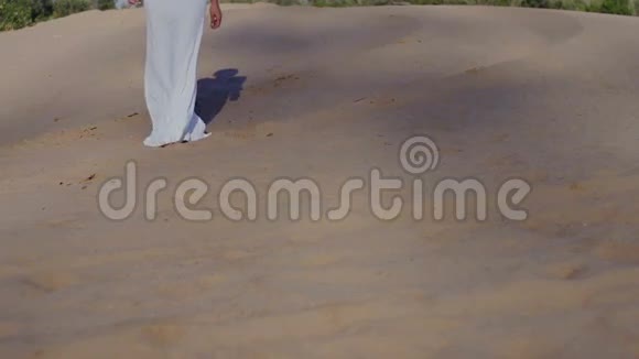 穿着白色连衣裙在海滩沙丘上行走的女人的脚太阳落山时的影子缓慢地向前看视频的预览图