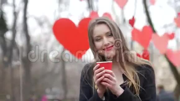 年轻美丽的金发女郎在秋天公园摆姿势装饰着红色的纸心背景和一杯视频的预览图